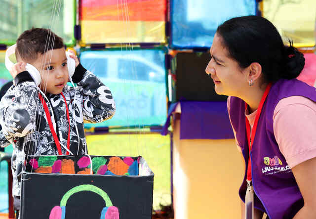 A three-year-old boy places headphones over his ears while participating in a JuJú a la Calle activity. Beside him, a member of the program staff leans down to speak with Alex.